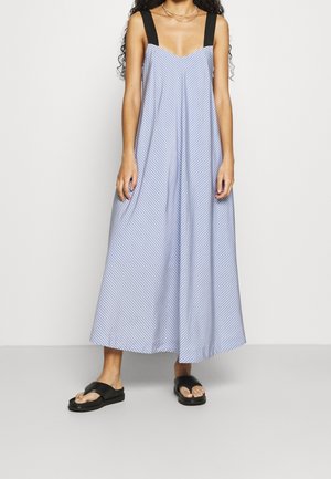 Vrouw draagt een mouwloze, wijd uitlopende jumpsuit met lichtblauwe en witte strepen, zwarte bandjes en zwarte sandalen.