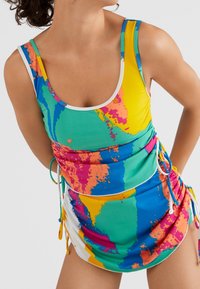 Femme portant un maillot de bain sans manches à motif abstrait multicolore avec des cordons latéraux, posant devant un fond clair.