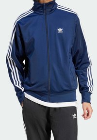 Marinblått zip-up träningsjacka med hög krage, vita tre-randsmönster på ärmarna och liten vit adidas-logotyp på bröstet.