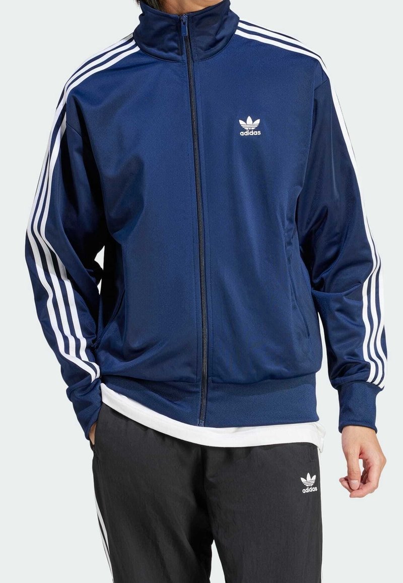 Marinblått zip-up träningsjacka med hög krage, vita tre-randsmönster på ärmarna och liten vit adidas-logotyp på bröstet.