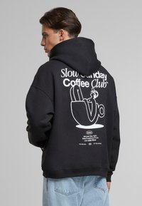 Black hoodie featuring a large white graphic print on the back with text and a coffee cup illustration. Soft fabric and relaxed fit.