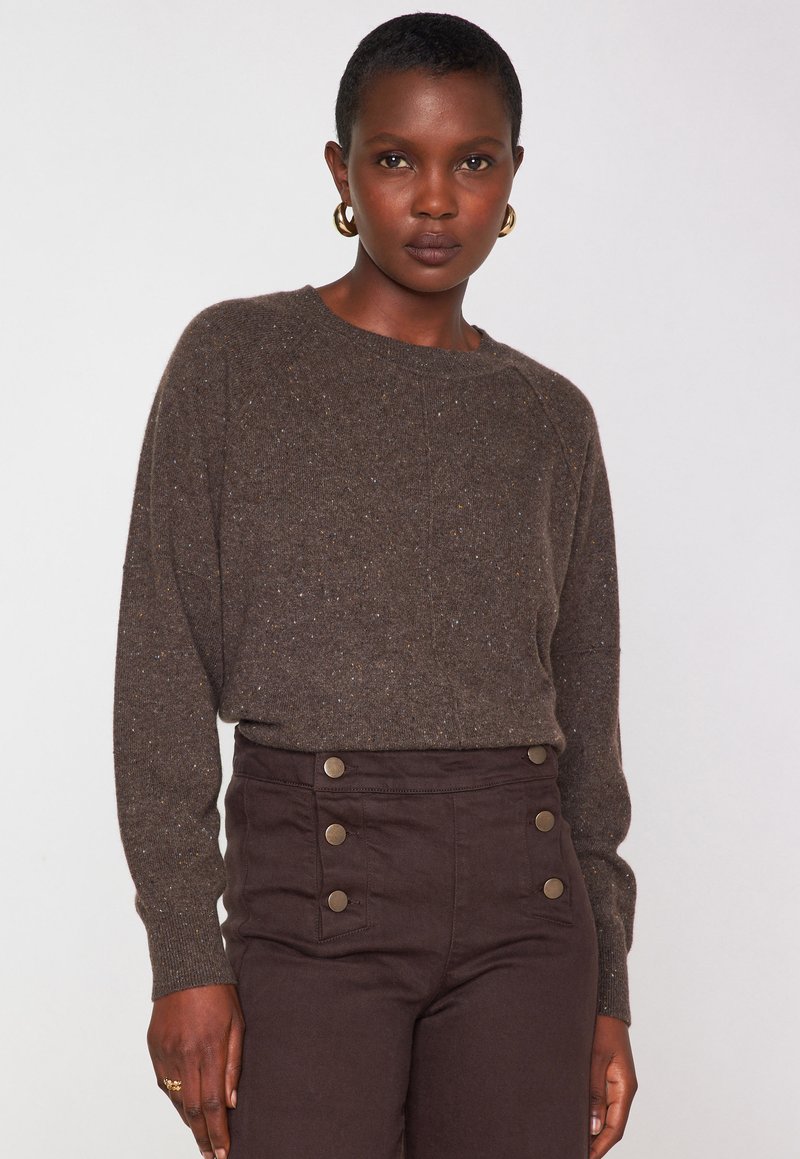 Femme aux cheveux courts portant un pull brun à taches et un pantalon brun taille haute avec six boutons en laiton, se tenant devant un fond uni.