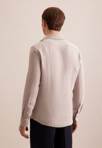 Camicia beige chiaro a maniche lunghe in un tessuto testurizzato, con colletto a punte e polsini con bottoni. La vista posteriore rivela una silhouette liscia.
