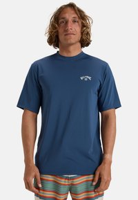 Rash guard de manga corta en azul marino con cuello redondo. Presenta texto de logo blanco en el pecho. Tejido suave y elástico.