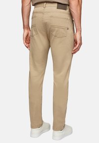 Beige cotton trousers with a straight cut, featuring two back pockets and a branded patch on the waistband. Worn with white sneakers.