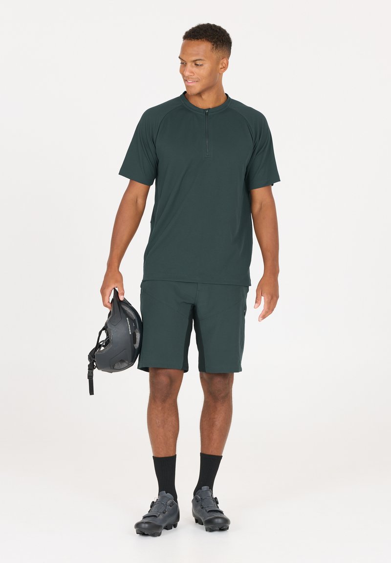Donkergroen sport t-shirt met korte mouwen en rits, bijpassende shorts, zwarte helm in de hand, zwarte sokken en fietsschoenen met een gestructureerde afwerking.