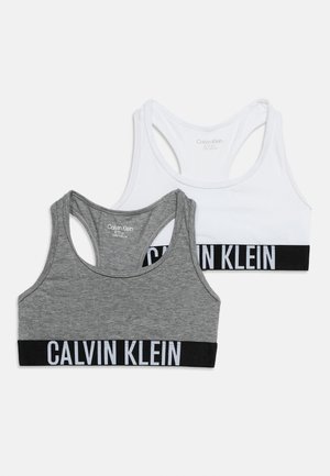 Két Calvin Klein márkájú sportmelltartó, az egyik fehér, a másik szürke, fekete elasztikus pántokkal, amelyeken fehér logós felirat található.