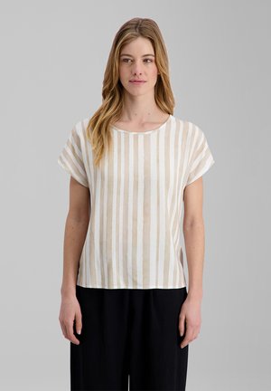 Frau mit langen blonden Haaren, die ein beige-weiß gestreiftes Kurzarmshirt und schwarze Hosen vor einem schlichten grauen Hintergrund trägt.