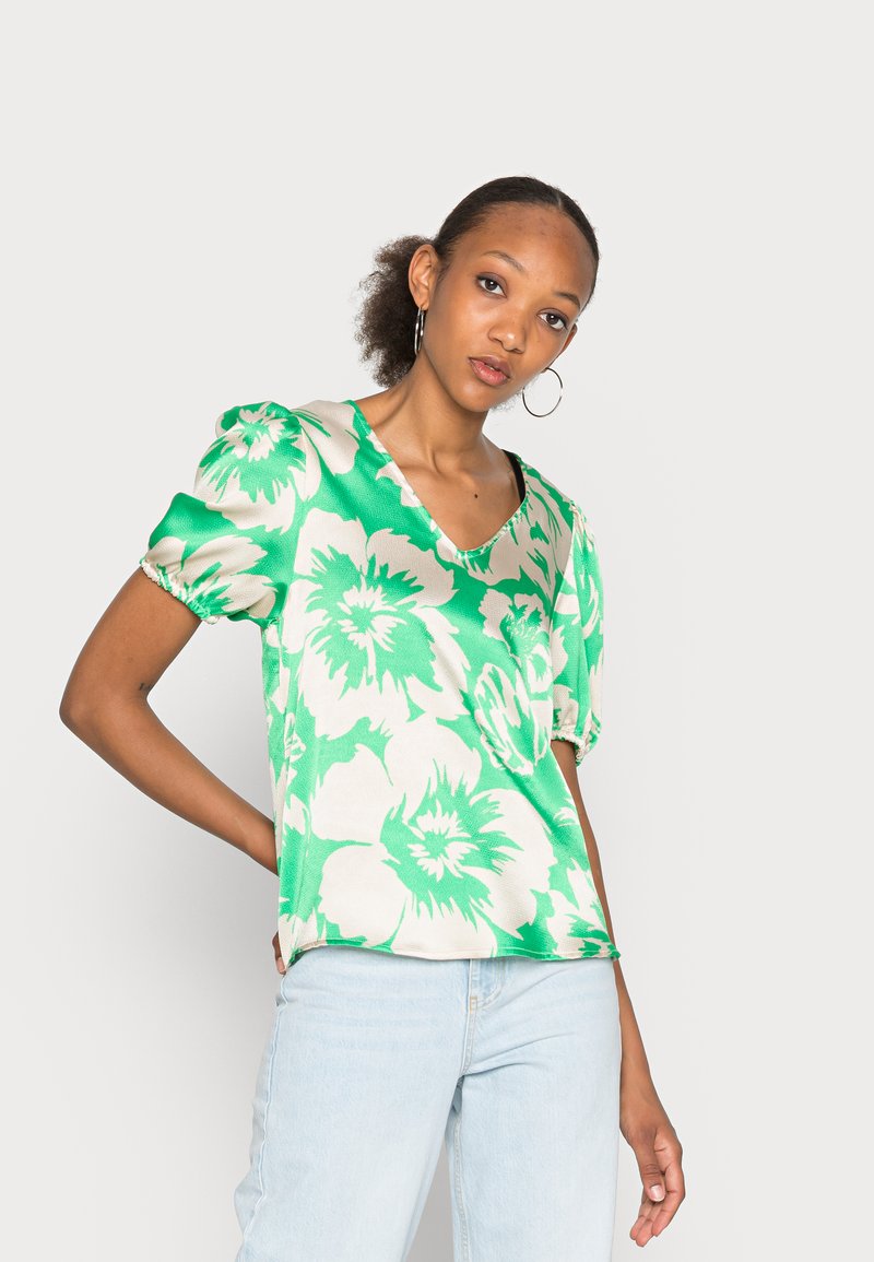 Top verde floral com mangas bufantes, decote em V e uma textura suave. O padrão apresenta grandes flores brancas sobre um fundo verde vibrante.