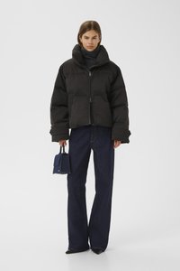 Svart pufferjakke med høy krage, teksturert overflate, glidelåslukking, båret over mørk denim flared bukser, kombinert med en liten blå håndveske.