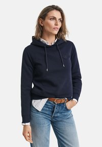 Marine hettegenser med snor, logodetalj og kengurulomme, lagd over en hvit stripete skjorte, parret med lyseblå jeans og brun belte.