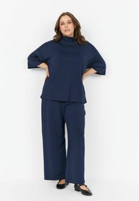 Femeie pozând într-un pulover larg de culoare albastră închis cu guler înalt și pantaloni largi, purtând pantofi plat negri, pe un fundal alb.