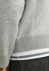 Grå ulltröja med ribbad nederkant, lager på lager över en vit skjorta med grå och blå ränder, tillsammans med mörk denimjeans.