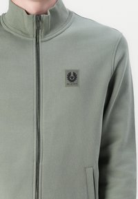 Gray-green zip-up jacket with high collar, textured fabric, side pockets, and a square black patch featuring a logo on the left chest.