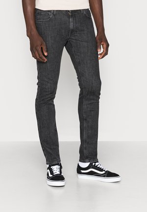 Person trägt schwarze Slim-Fit-Jeans und schwarz-weiße Sneakers und steht vor einem schlichten Hintergrund.