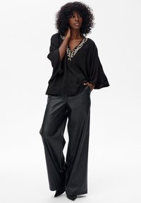 Schwarze, fließende Bluse mit besticktem V-Ausschnitt; kombiniert mit weiten, schwarzen Lederhosen. Beide Teile haben eine glatte Textur.