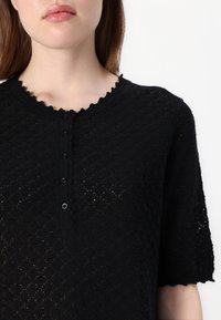 Top nero a maglia con maniche corte, caratterizzato da un colletto a onde e una fila di bottoni, che mostra un motivo testurizzato su tutta la superficie.