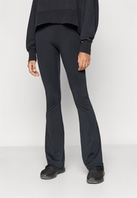 Μαύρα κολάν με φαρδύ καμπάνα, κατασκευασμένα από λείο, ελαστικό υλικό. Διαθέτουν ψηλή μέση και ελαφριά γυαλάδα, συνδυασμένα με μαύρα αθλητικά παπούτσια.