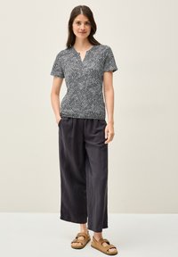 Blouse noire et blanche à manches courtes avec des motifs ondulés, associée à un pantalon noir ample et des sandales marron. Coupe décontractée et tissu léger.