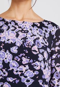 Zbliżenie osoby w czarnej bluzce z abstrakcyjnym wzorem kwiatowym w odcieniach fioletu i różu, ukazujące dekolt i strukturę tkaniny.