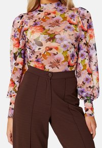 Blommönstrad långärmad topp med hög hals, genomskinligt tyg med rosa, orange och lila blommor, kombinerad med bruna tajta byxor.