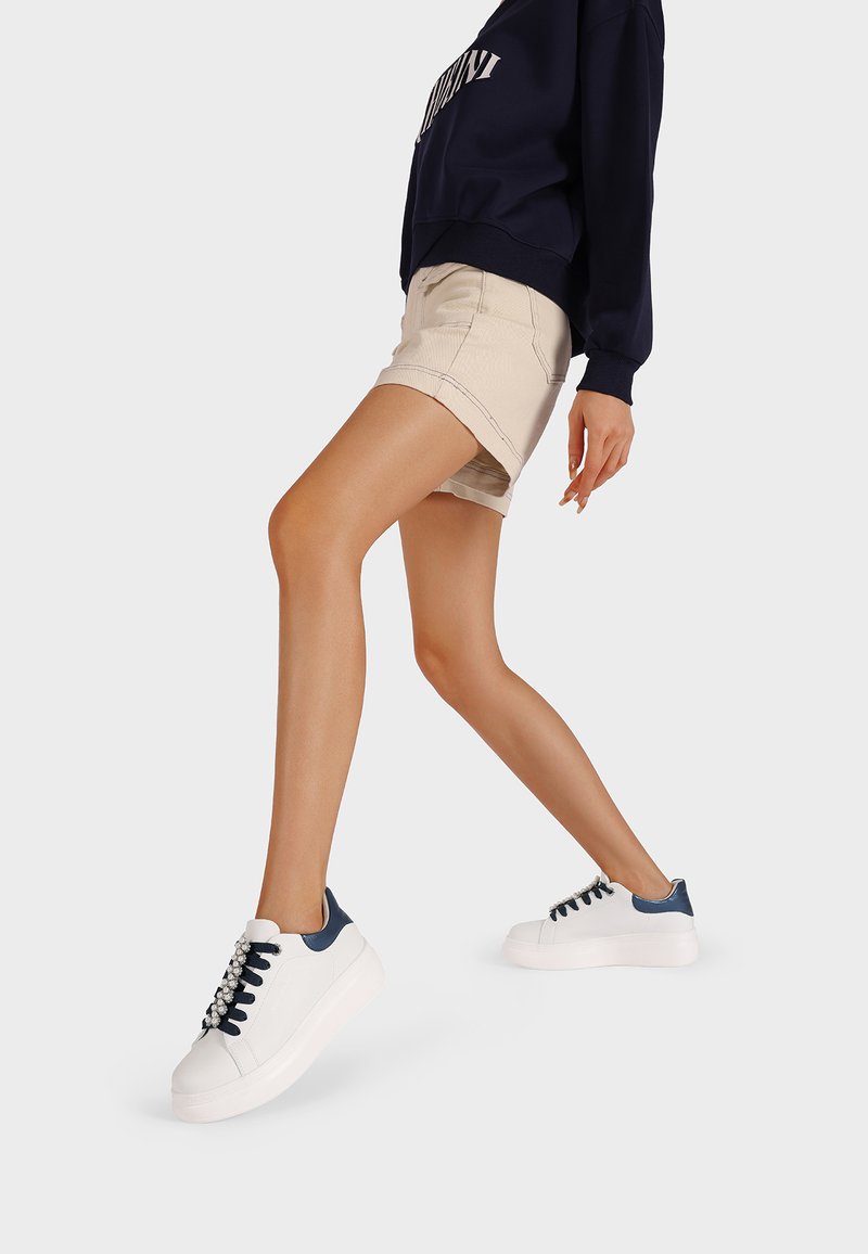 Sneaker bianco con accenti navy, texture in pelle liscia, punta rotonda e lacci decorativi. Indossate con pantaloni corti beige e una felpa navy.
