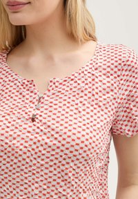 Blouse à motifs rouges et blancs avec un décolleté arrondi, des boutons décoratifs et des manches courtes froncées. Tissu léger avec une finition texturée.