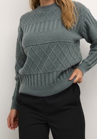 Donna che indossa un maglione grigio a maglia con motivi a diamante e a coste, abbinato a pantaloni neri, in piedi davanti a uno sfondo chiaro e uniforme.