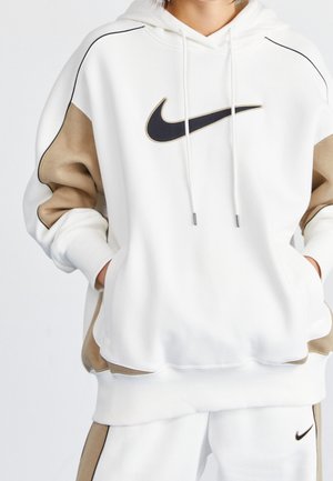 Personne portant un sweat à capuche Nike blanc et beige et un pantalon de survêtement assorti, mains dans la poche avant, grand logo Nike noir visible sur la poitrine.