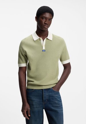 Junger Mann trägt ein hellgrünes kurzärmeliges Strick-Poloshirt mit weißem Kragen und Manschetten und blaue Jeans, steht mit einer Hand in der Tasche vor weißem Hintergrund.