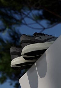 Gray and black Adidas sneakers with white soles resting on a white surface against a blurred blue sky and tree background.