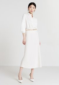 Blusa bianca a maniche lunghe con colletto, abbinata a una gonna midi crema. Vita accentuata da una sottile cintura dorata e indossata con tacchi bianchi a punta.