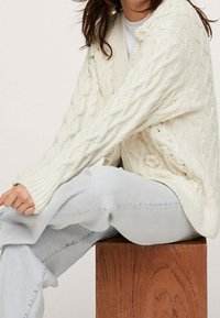 Woman wearing a cream cable-knit cardigan and light blue jeans, sitting sideways on a wooden cube against a neutral background.