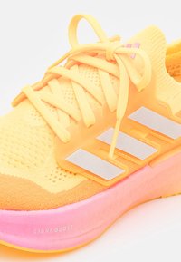 Chaussure de course jaune en maille avec semelle rose, trois bandes réfléchissantes sur le côté et lacets jaunes attachés en nœud.