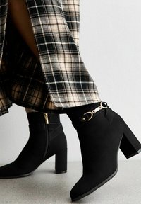 Botas negras hasta el tobillo de ante con un tacón cuadrado. Incluyen una correa decorativa y detalles en dorado. Combinadas con una falda de cuadros.