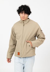 Beżowa, lekka kurtka z wysokim kołnierzem, zamkiem błyskawicznym, bocznymi kieszeniami i pomarańczową metką na dolnym brzegu. Charakteryzuje się teksturowanym, gładkim wykończeniem.