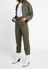 Olivgrön jacka med knappstängning och bröstficka, ihopparad med matchande byxor och vita ankelstövlar med fyrkantig klack.