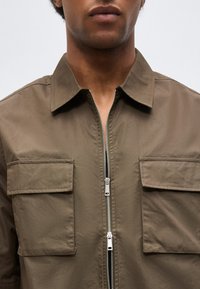 Κοντινό πλάνο ενός ατόμου που φορά ένα καφέ πουκάμισο-ζακέτα με δύο εμπρόσθιες τσέπες με καπάκι και ένα ασημένιο διπλό φερμουάρ, μερικώς ξεκουμπωμένο.