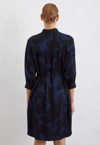 Robe bleu marine en tissu lisse avec un motif abstrait sombre, des manches à boutons et une taille cintrée pour une définition de la silhouette.