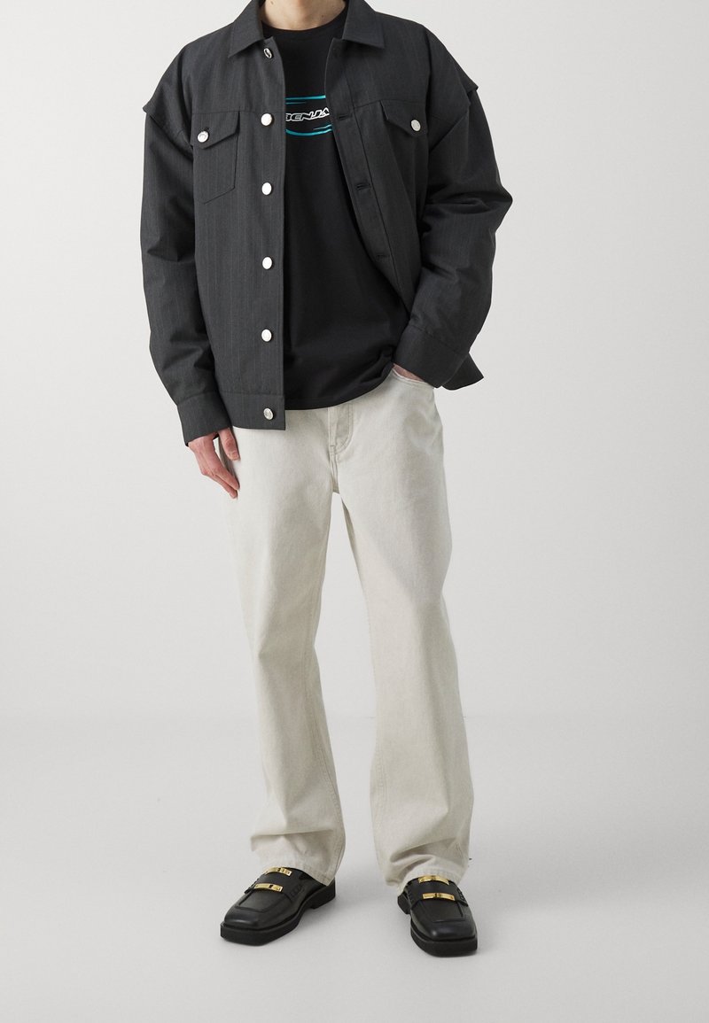 Svart kavaj med smala ränder, svart skjorta med logotyp, ljusbeiga byxor och svarta loafers med gulddetaljer. Fokusera på fickor och knappar.