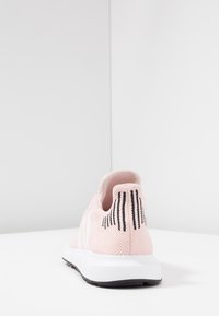 Vaaleanpunainen neulottu urheilukenkä, jossa on teksturoitu päällinen, valkoinen välipohja ja mustat raidalliset yksityiskohdat kantapäässä. Kevyt ja hengittävä muotoilu.