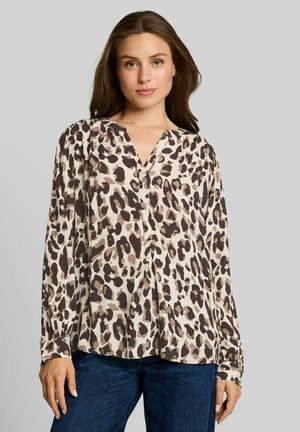 Frau mit langen braunen Haaren, die eine beige und braune Leopardenmuster-Bluse und dunkelblaue Jeans trägt, steht vor einem neutralen Hintergrund.