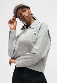 Person adjusting beige cap, wearing a gray and white striped button-up shirt with Carhartt logo and black pants on a plain background.