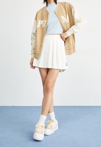 Tan- en crèmekleurig varsityjack met witte letters, lichtblauw topje, witte geplooide rok en stevige witte sneakers met beige accenten.