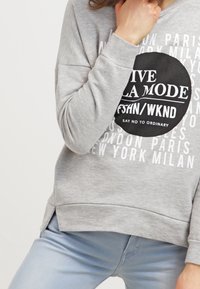 Sudadera gris con un gráfico redondo negro y texto blanco. Tiene un corte relajado, mangas largas y un dobladillo redondeado.