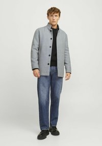 Abrigo de mezcla de lana gris con cuello alto, cierre de botones y bolsillos laterales, llevado sobre un suéter negro y jeans azules. Zapatos negros completan el conjunto.