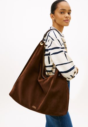 Γυναίκα που φοράει λευκό πουκάμισο με ναυτικές αφηρημένες γραμμές και μπλε τζιν, κρατώντας μεγάλη καφέ σουέντ τσάντα ώμου με χρυσά μεταλλικά στοιχεία.