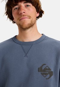 Felpa blu in cotone, con collo a giro, polsini a costine e un logo nero sul lato sinistro. Texture liscia e design semplice.