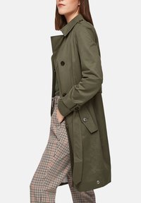 Trench coat verde oliva con bottoni doppi, tasche laterali e vita con cintura. Il tessuto ha una consistenza liscia, adatta per essere indossata a strati.