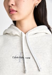 Sweatshirt Calvin Klein em cinza claro, feito de um tecido suave com um bolso frontal. Apresenta um capuz com cordão e logótipo bordado em cores contrastantes.